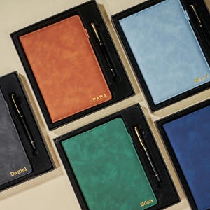 May include: A selection of leather notebooks in different colours, each accompanied by a black pen, presented in individual black boxes. The notebooks are in shades of orange, blue, green, grey, and navy. Some notebooks have gold lettering with names.