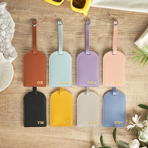 Könnte beinhalten: Acht farbige Gepäckanhänger in verschiedenen Farbtönen, darunter Braun, Blau und Gelb, sind auf einer Holzoberfläche angeordnet. Jeder Anhänger hat eine goldfarbene Initialen oder einen Namen. Die Anhänger sind rechteckig mit abgerundeter Oberseite und einem Riemen.