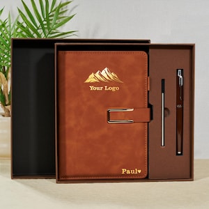 May include: A brown leather journal with a gold mountain logo and the text "Your Logo". The journal has a metal clasp and the name "Paul" with a heart symbol. A pen and refill are included in the box.