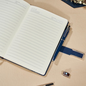 May include: An open, lined notebook with a dark blue cover and a matching pen. The pen is secured by a blue strap with a gold-colored accent. A small, decorative object with a blue gem sits nearby.