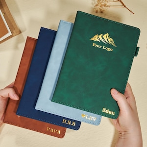 Op de afbeelding: Een verzameling gepersonaliseerde notitieboekjes in verschillende kleuren: bruin, marineblauw, lichtblauw en groen. Het groene notitieboekje heeft een gouden berglogo met de tekst "Your Logo" en "Eden". Andere notitieboekjes hebben gouden letters met namen of initialen.
