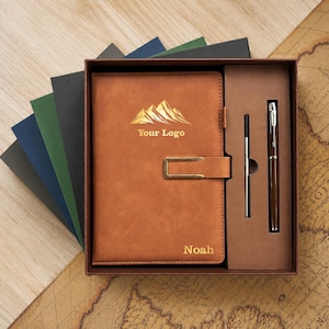 Peut inclure: Un carnet marron clair avec un motif de montagne doré, le texte "Your Logo" et le nom "Noah" gravés sur la couverture. Le carnet possède une attache métallique dorée et est présenté dans une boîte avec un stylo assorti et un stylet fin.