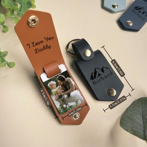 May include: A brown leather keychain with a photo insert and a message that says "I Love You Daddy". The keychain also has a black leather tag with a mountain design and the name "Richard" engraved on it. The keychain measures 1.7 inches by 2.6 inches.