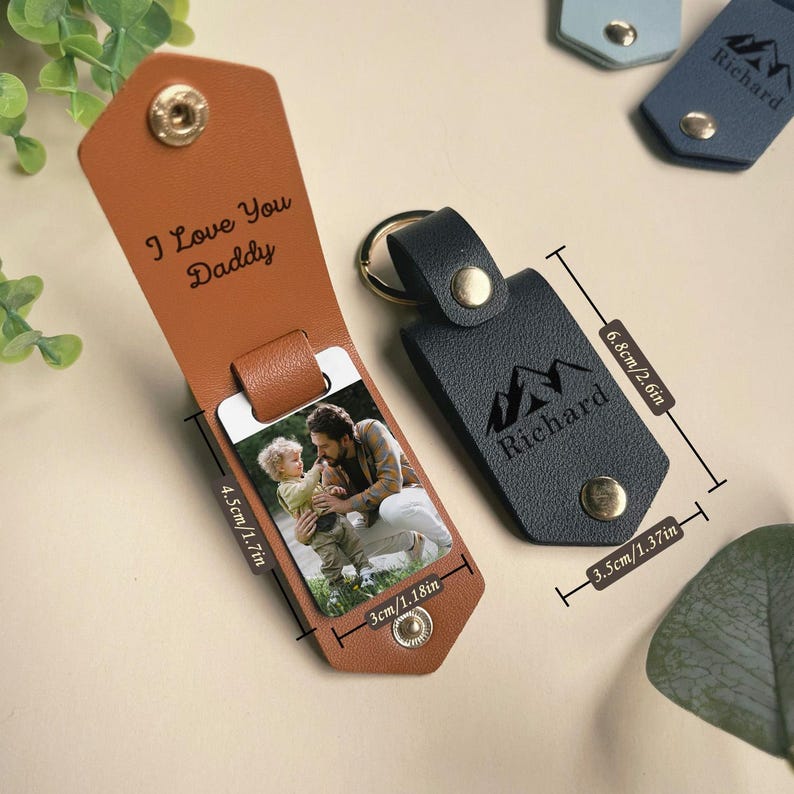 May include: A brown leather keychain with a photo insert and a message that says "I Love You Daddy". The keychain also has a black leather tag with a mountain design and the name "Richard" engraved on it. The keychain measures 1.7 inches by 2.6 inches.