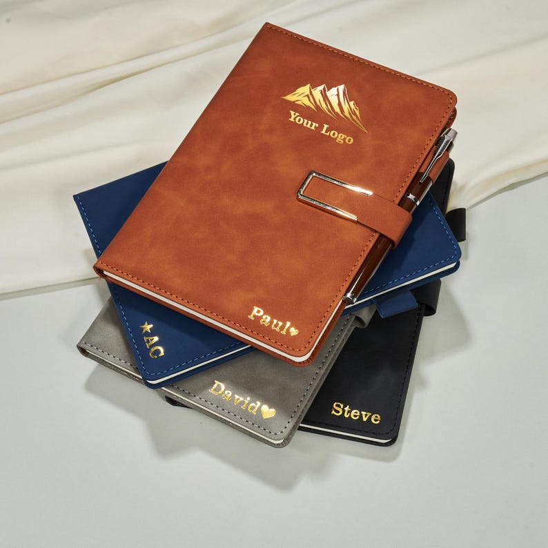 May include: A stack of personalized notebooks in various colors, including brown, navy, gray, and black. The brown notebook features a gold mountain logo and the text "Your Logo" and "Paul". Each notebook has a pen holder and a silver clasp.