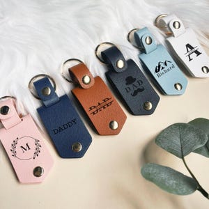 May include: Six leather keychains with different designs and engravings. The keychains are in various colors, including brown, blue, pink, and light blue. The engravings include "DAD", "Daddy", "M", "Richard", and "Aiden".
