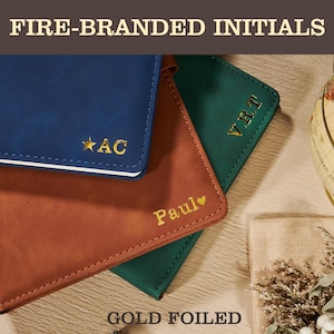 May include: A close-up of three personalized notebooks in blue, brown, and green. Each notebook features gold foil initials or a name with a heart symbol. The text "FIRE-BRANDED INITIALS" and "GOLD FOILED" are at the top and bottom.