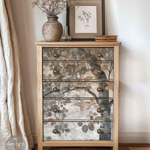 May include: A five-drawer wooden chest with a tree and leaf design in grey, brown, and beige. The chest has brass knobs and is topped with decorative items, including a framed botanical print and a vase. The chest is made of wood.