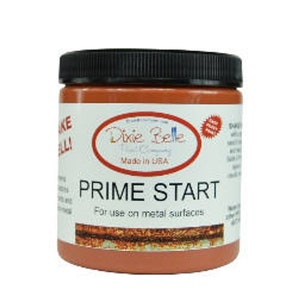 Op de afbeelding: Een pot Dixie Belle Paint Company Prime Start primer voor metalen oppervlakken. De pot is gelabeld met het logo van het bedrijf en de productnaam. De pot is roestoranje van kleur.