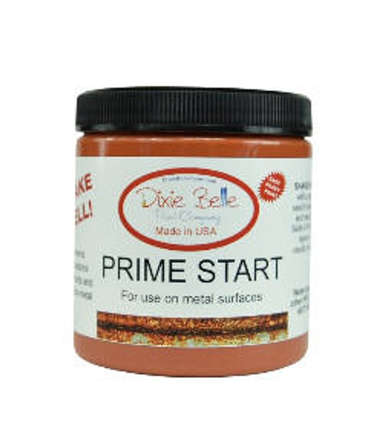 Op de afbeelding: Een pot Dixie Belle Paint Company Prime Start primer voor metalen oppervlakken. De pot is gelabeld met het logo van het bedrijf en de productnaam. De pot is roestoranje van kleur.