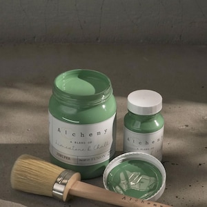 May include: Two bottles of green paint labeled "Alchemy, A Blend of Limestone & Chalk" and "Juniper, Paint by Fusion". A paint brush with a wooden handle is lying on a concrete surface.