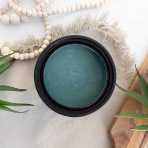 May include: A teal-coloured paint tin with a black rim, surrounded by decorative items. A wooden chopping board, faux succulents, and a strand of wooden beads are arranged around the paint tin on a neutral-toned surface.
