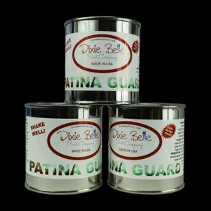 Op de afbeelding: Drie zilveren blikken Dixie Belle Patina Guard verf. De blikken zijn gelabeld met het Dixie Belle logo en de woorden "Patina Guard" in rood en groene letters. De blikken zijn op elkaar gestapeld.