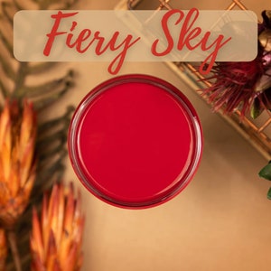 May include: A small jar of red paint with the text "Fiery Sky" above it. The jar is on a brown surface with dried flowers and greenery around it.