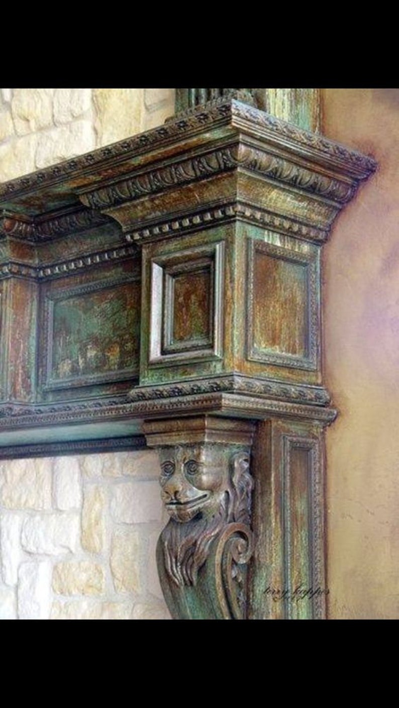 Op de afbeelding: Een close-up van een decoratieve schouw met ingewikkelde houtsnijwerk en een groene en bruine patina. De schouw heeft een gegraveerd leeuwenhoofd in de rechterbenedenhoek.
