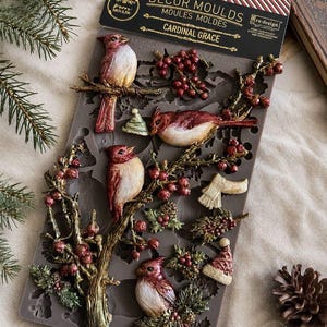 May include: A grey silicone decor mould titled Cardinal Grace featuring recessed designs of four red cardinals perched on branches with holly berries, pine cones, a small hat, and a scarf. The packaging header is black with white and gold text.