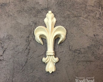 May include: A light-colored, fleur-de-lis shaped wooden decorative element. The design features three prominent petals at the top, a central stem, and a stylized base. The background is a textured, dark gray surface.