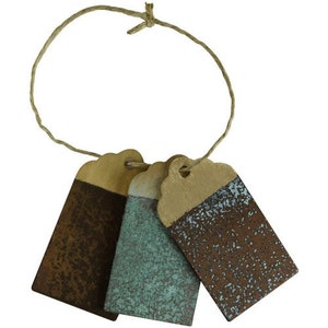 Op de afbeelding: Drie rustieke, rechthoekige, beschilderde houten labels met een touwtje. De labels zijn geschilderd in tinten bruin, teal en blauw met een gespikkelde afwerking.