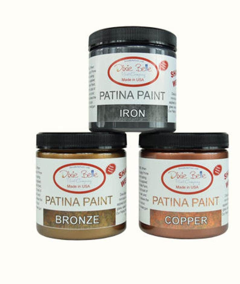 Op de afbeelding: Drie glazen potten met Dixie Belle Patina-verf in de kleuren ijzer, brons en koper. Elke pot heeft een zwart deksel en een wit etiket met het Dixie Belle-logo en de woorden "Patina Paint" en de kleurnaam.