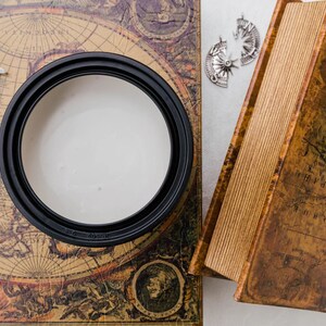 May include: A black candle holder with a white candle inside, sitting on a vintage map with a silver compass charm and a crescent moon charm.