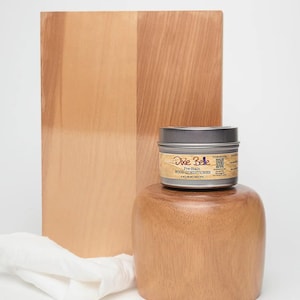 May include: A wooden chopping board and a wooden pedestal with a small metal tin of Dixie Belle Pre-Stain Wood Conditioner. A white cloth is draped on the surface. The wood has a light brown colour.