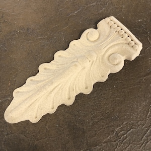 May include: A beige, ornate, wooden decorative trim with a leaf-like design. The trim has a raised, textured surface and a beaded edge.