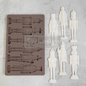 May include: A silicone mould with eight cavities shaped like nutcracker figurines. The mould is brown and the figurines are white. The mould is labelled "La Design with Prima" and "www.ladesignwithprima.com".