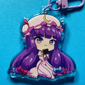 Könnte beinhalten: Ein lila und pinkfarbener Anime-Charakter-Schlüsselanhänger mit einem Halbmond auf ihrem Hut. Sie hält eine schwarze Karte in der Hand.