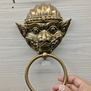 Puede incluir: Un toallero de pared de color latón con una cara estilizada detallada y un anillo circular. La cara tiene una expresión sonriente y está montada sobre una superficie de madera de color claro.