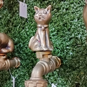 May include: A brass garden hose faucet with a cat figurine on top. The cat is sitting upright with its paws together and has a surprised expression on its face. The faucet is a traditional style with a round handle and a spout that curves downwards.