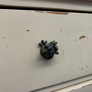 May include: A close-up of a white painted drawer with a frog-shaped knob. The knob is dark blue and gold, and the drawer has a distressed, crackled paint finish. A gold knob is visible to the right.