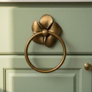 May include: A bronze-colored flower-shaped cabinet pull with a circular handle. The hardware is mounted on a light green cabinet door with a matching round knob. The design is a decorative accent for furniture.