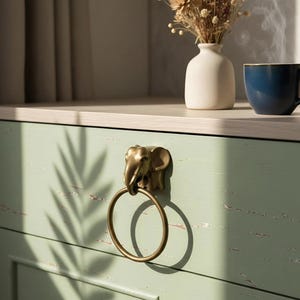 Könnte beinhalten: Ein Schubladengriff in Form eines Elefanten aus Messing mit einem Ringgriff ist an einer hellgrünen Schranktür angebracht. Eine kleine weiße Vase mit Trockenblumen und eine blaue Tasse mit Dampf stehen auf der Arbeitsplatte.