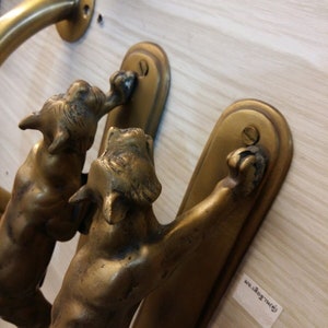 May include: Two brass door handles featuring a cat design. The cats are depicted in a leaping pose with their paws raised. The handles are mounted on a wooden surface.