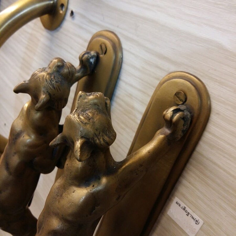 May include: Two brass door handles featuring a cat design. The cats are depicted in a leaping pose with their paws raised. The handles are mounted on a wooden surface.