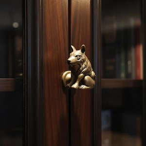 May include: A bronze cabinet door pull in the shape of a seated wolf. The hardware is mounted on a dark wood cabinet door. The cabinet door has a rich, dark brown colour with visible wood grain.