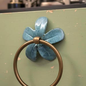 May include: A drawer pull with a blue flower design and a brass ring handle. The flower has a textured finish and is mounted on a distressed green surface. The hardware adds a touch of vintage charm and is suitable for a cabinet.