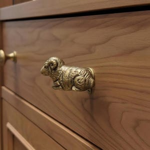 Könnte beinhalten: Ein bronzefarbener Schubladengriff in Form eines Widders, der an einem Holzschrank befestigt ist. Der Widder ist mit gekräuselten Hörnern und strukturiertem Fell detailliert. Der Schrank hat eine natürliche Holzmaserung.