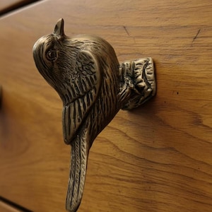 May include: A bronze-colored bird-shaped drawer pull. The pull is attached to a wooden drawer. The bird has detailed feathers and a curved beak. The drawer is made of light brown wood.
