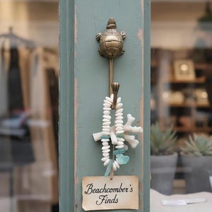 May include: A bronze turtle-shaped hook hangs on a weathered, teal-painted wooden post. Below the hook, a decorative strand of white beads, coral, and sea glass is suspended. A small tag reads "Beachcomber's Finds."