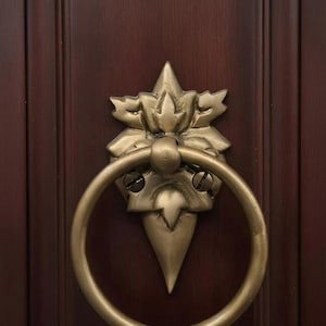 Op de afbeelding: Een sierlijke messing deurklopper met een ringhandvat en een decoratief bloemmotief. De klopper is gemonteerd op een donkerbruine houten deur, wat een klassieke en elegante uitstraling creëert.