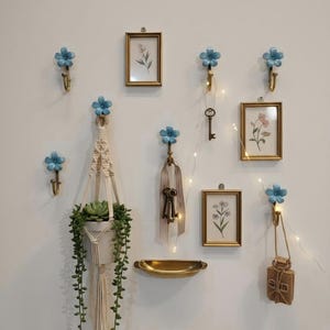 May include: A wall display featuring blue flower-shaped hooks, small framed botanical prints, and a macrame plant hanger with a trailing succulent. Decorative keys and a small bag hang from the hooks. A brass-toned shelf is also included.