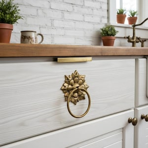 Puede incluir: Primer plano de un mueble de cocina blanco con un tirador de anillo floral dorado y una manija de barra dorada. El mueble forma parte de una cocina con encimera de madera y pared de ladrillo blanco. Se ven pequeñas plantas en macetas.