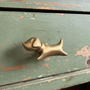 Op de afbeelding: Een messingkleurige hondenknop voor een lade. De hond is afgebeeld in een speelse pose, met zijn staart omhoog en kop naar voren. Gemonteerd op een verweerde, groen geschilderde houten lade.