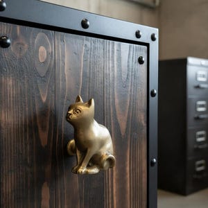 Pode incluir: Um puxador de armário em forma de gato de latão está montado numa porta de armário de madeira escura. A porta é emoldurada com uma borda de metal preto e rebites. O armário está numa sala com um fundo neutro.
