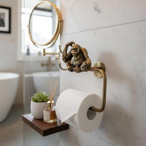 Könnte beinhalten: Ein dekorativer Toilettenpapierhalter in Bronzeoptik mit einer Orang-Utan-Skulptur. Der Halter ist an einer Wand mit Marmormuster befestigt und hält eine Rolle weißes Toilettenpapier. Ein goldfarbener Spiegel und eine Ablage mit Zubehör sind ebenfalls zu sehen.