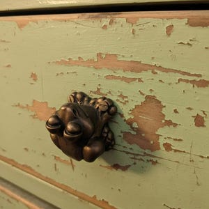 Op de afbeelding: Close-up van een bronzen ladeknop met een gedetailleerd, sierlijk ontwerp. De knop is bevestigd op een verweerde, lichtgroen geschilderde houten lade, met zichtbare slijtage en blootliggend hout.