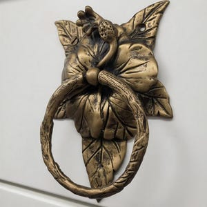 Op de afbeelding: Bronzen deurklopper in de vorm van een bloem met bladeren. De klopper heeft een ringvormige handgreep en is op een wit oppervlak gemonteerd. Het bloemmotief voegt een decoratief element toe aan elke deur.