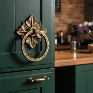 Puede incluir: Primer plano de un tirador de puerta de armario de latón con diseño floral y asa de anillo. La ferretería está montada en una puerta de armario verde oscuro. Se ve un tirador a juego debajo. El fondo muestra una cocina.