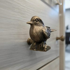 Puede incluir: Un pomo de armario con forma de pájaro de color bronce. El pomo está montado en un armario de madera de color claro. El pájaro está posado en una pequeña rama, con plumas detalladas y un aspecto realista. Se ven otros pomos a juego.